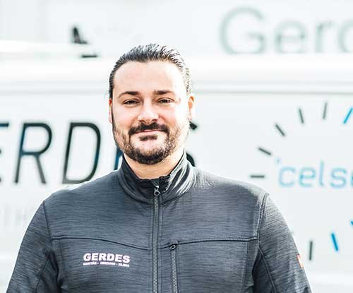 Ein Mann mit einem Bart trägt eine dunkle Jacke und lächelt vor einem Fahrzeug mit dem Schriftzug 'GERDES'.