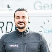 Professioneller Handwerker vor einem Firmenfahrzeug Ein Mann mit einem Bart trägt eine dunkle Jacke und lächelt vor einem Fahrzeug mit dem Schriftzug 'GERDES'.