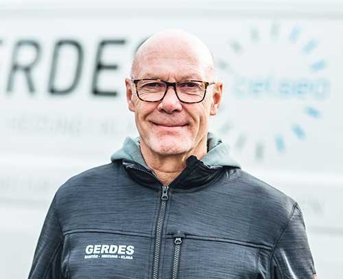 Ein älterer Mann mit Brille und Glatze lächelt freundlich in die Kamera, während er einen schwarzen Jacke trägt.