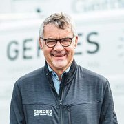 Lächelnder Mann in moderner Arbeitsumgebung Ein lächelnder Mann mit Brille trägt eine dunkle Jacke und steht vor einem hellen Hintergrund mit dem Schriftzug 'GERDES'.