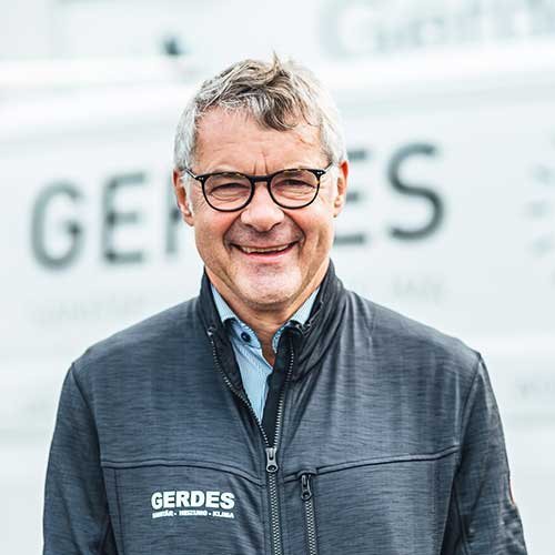 Ein lächelnder Mann mit Brille trägt eine dunkle Jacke und steht vor einem hellen Hintergrund mit dem Schriftzug 'GERDES'.