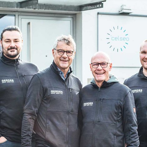 Vier Männer in schwarzen Jacken stehen lächelnd vor einem Firmengebäude mit dem Namen 'Gerdes'.